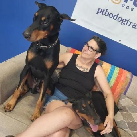 ineke and dogs tilt Ineke sitting on a couch with two Dobermans—one sitting tall and alert, the other resting across her lap—at Turner & Pooch Dog Training.